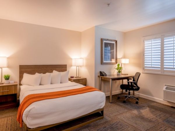 The image shows a cozy hotel room with a double bed, orange throw blanket, two bedside tables, a desk, chair, and window with blinds.