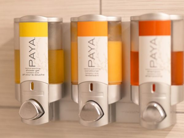 The image shows three wall-mounted dispensers labeled "PAYA" with various colors of liquid, likely shower gel or soap, in a bathroom setting.