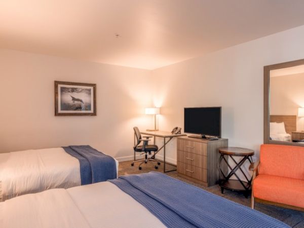 The image shows a hotel room with two beds, a TV, desk, chair, lamp, and wall art. There's a mirror and an orange chair next to the desk.