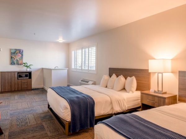 The image shows a hotel room with two double beds, a lamp, a small kitchenette, and a piece of artwork on the wall.