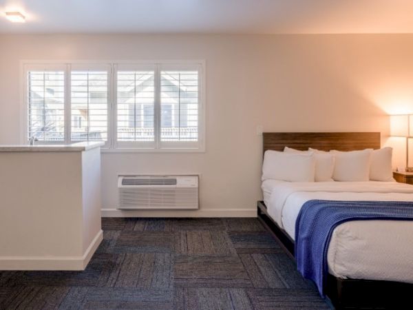 The image shows a hotel room with a bed, bedside lamp, window with shutters, air conditioning unit, and partial wall.