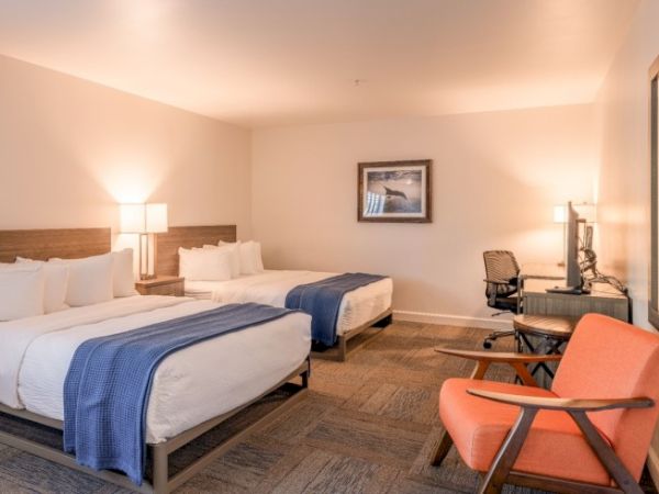 A hotel room with two double beds, a desk with chair, orange armchair, lamps, and artwork on the wall, looking clean and cozy.