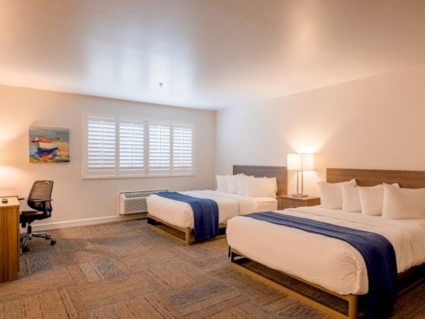 A hotel room with two beds, a desk with a chair, an artwork on the wall, and a lamp, all under a bright ceiling light.