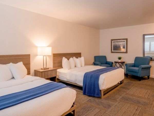 A hotel room with two double beds, blue accents, a lamp, artwork, two chairs, and a small table, creating a cozy, inviting atmosphere.