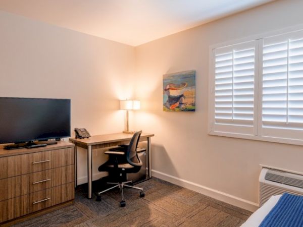 The image shows a hotel room with a desk, chair, TV, dresser, lamp, and a window with shutters. There's a painting on the wall.