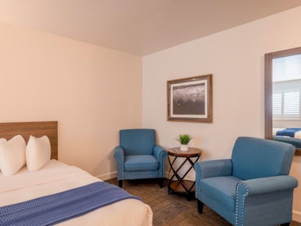 The image shows a cozy room with a bed, two blue armchairs, a small round table, and a mirror on the wall.