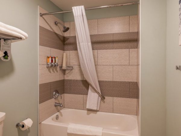 The image shows a bathroom with a bathtub, showerhead, curtain, and toiletries on the wall. Towels are neatly folded on a rack.