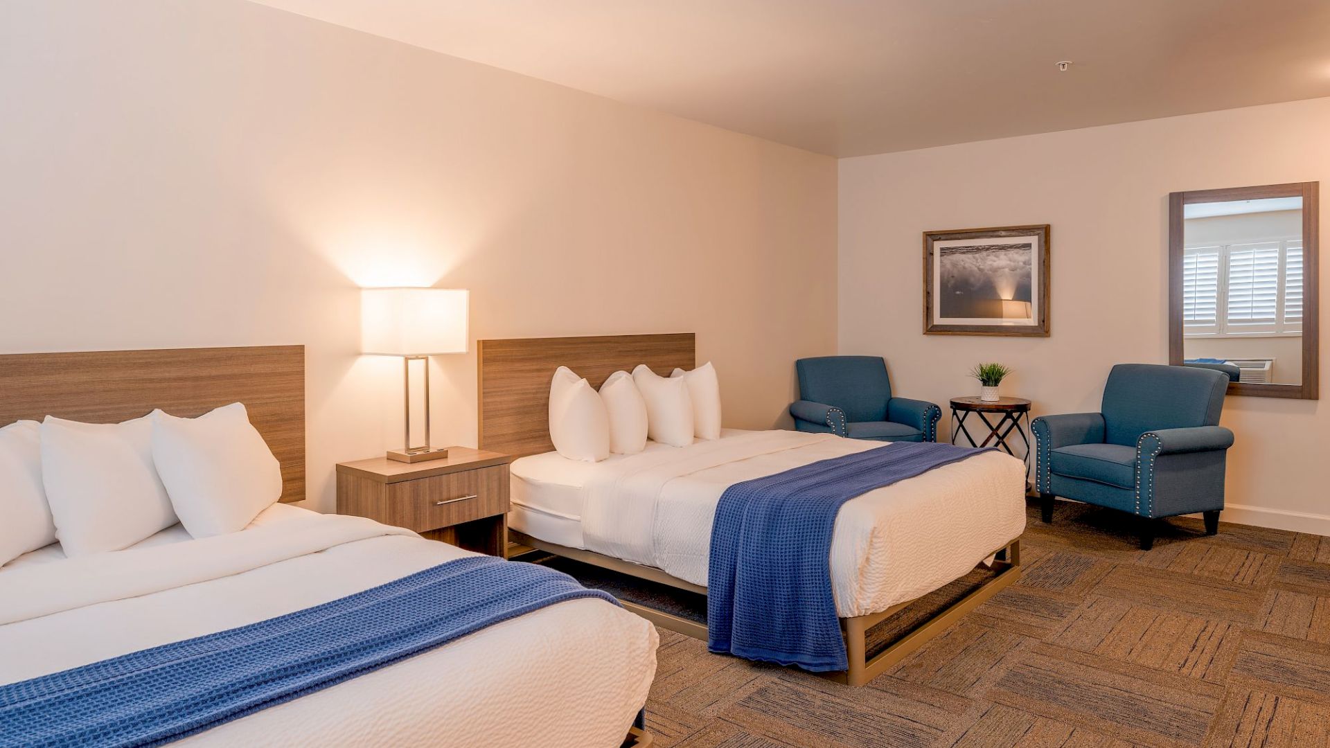 A hotel room with two beds, blue blankets, a lamp, chairs, a side table, and a large mirror on the wall.