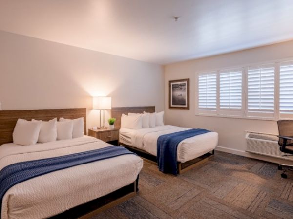 The image shows a hotel room with two beds, blue throws, a lamp on a nightstand, a chair, window shutters, and a wall picture.