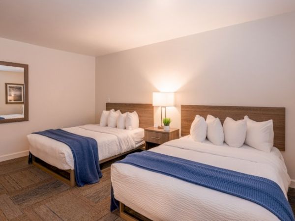 The image shows a hotel room with two double beds, white linens, blue throws, a bedside table, a lamp, and a mirror on the wall.