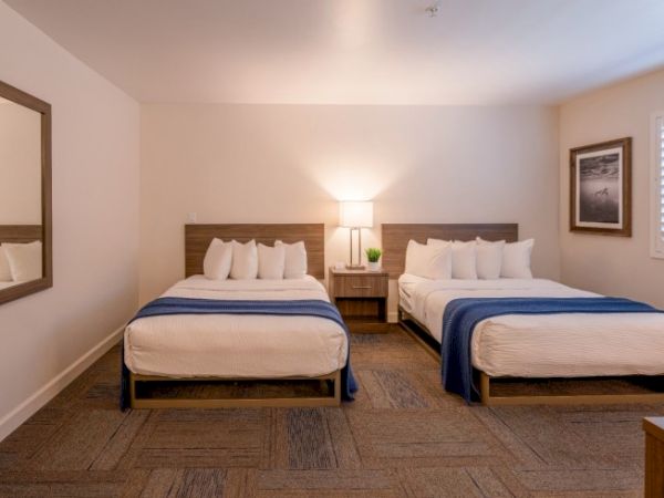 The image shows a hotel room with two neatly made beds, a nightstand with a lamp, a mirror, and a framed picture on the wall.