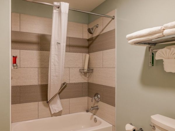 The image shows a bathroom with a bathtub, shower curtain, tiled walls, towels on a rack, and a towel ring with a decorative folded towel.