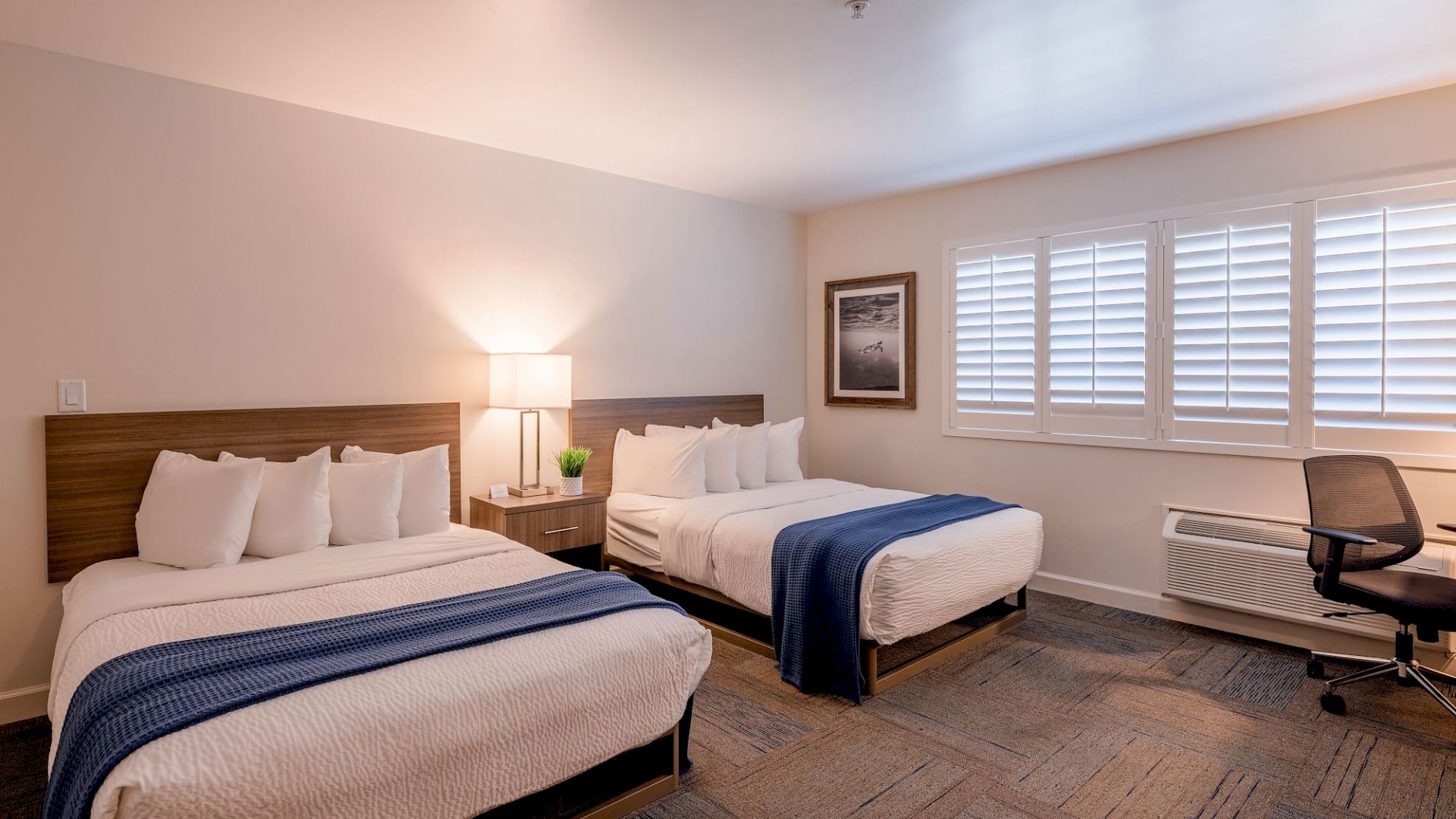 The image shows a hotel room with two beds, each with a blue throw, a lamp, a desk with a chair, and a window with shutters.