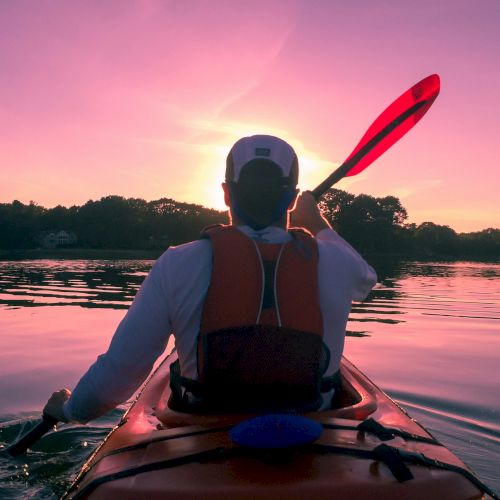 A person paddling a red kayak on calm water at sunset, wearing a life jacket and holding a paddle over their shoulder.