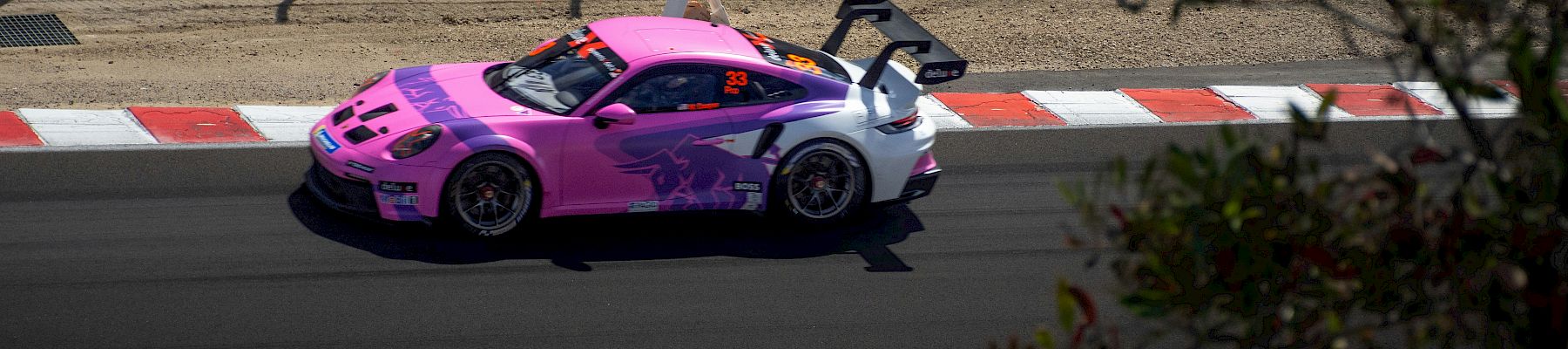 A pink-and-purple race car with the number 33 speeds on a track behind fencing and lane markings, under sunny conditions.