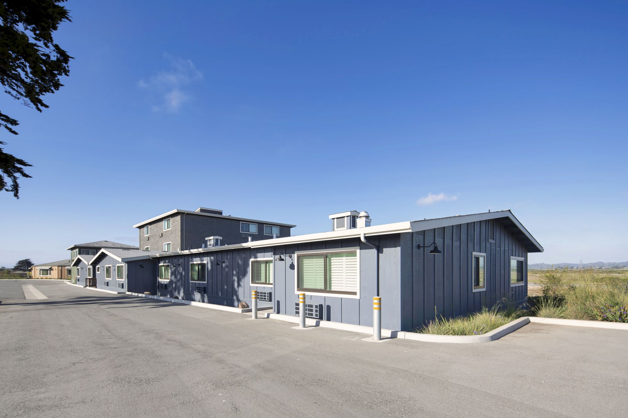 The image shows a row of modern, single-story buildings with a paved road, clear skies, and grassy areas in the background.