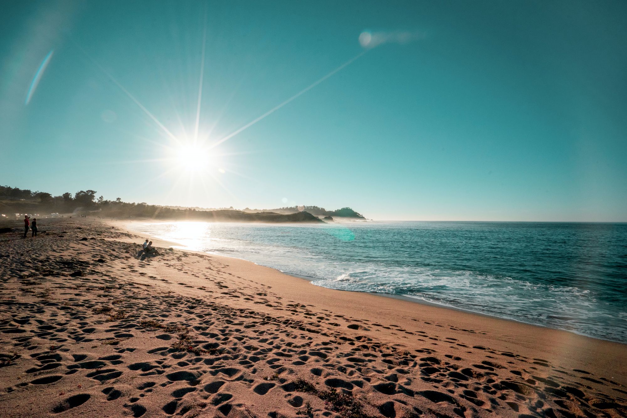 A sunlit beach with footprints in the sand, waves gently crashing, clear blue sky, and distant vegetation and hills on the horizon.