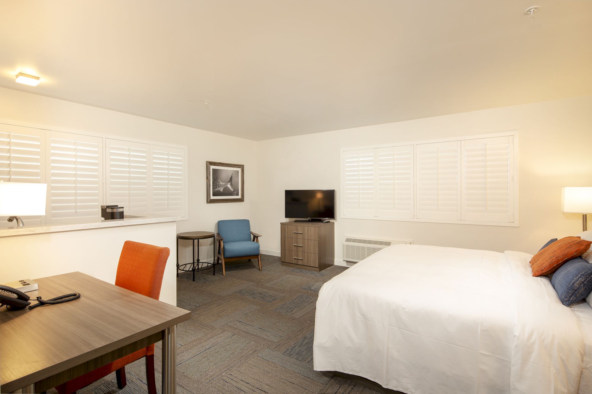 A spacious, well-lit hotel room with a bed, desk, TV, chairs, and a dresser. The room features a large window with shutters and modern decor.