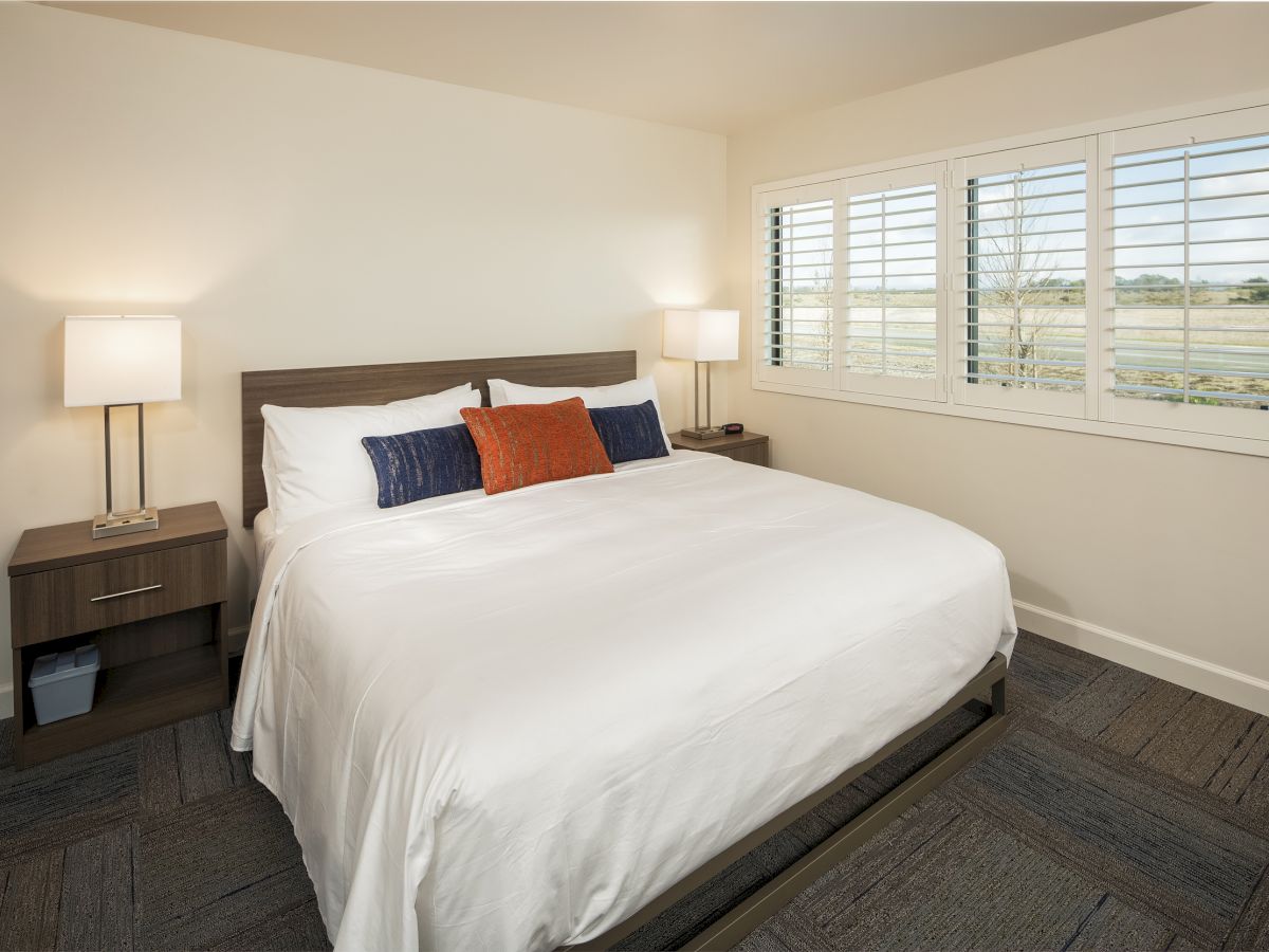 A neatly arranged bedroom with a large bed, white bedding, and colorful pillows. There are two bedside tables with lamps, and a window with shutters.