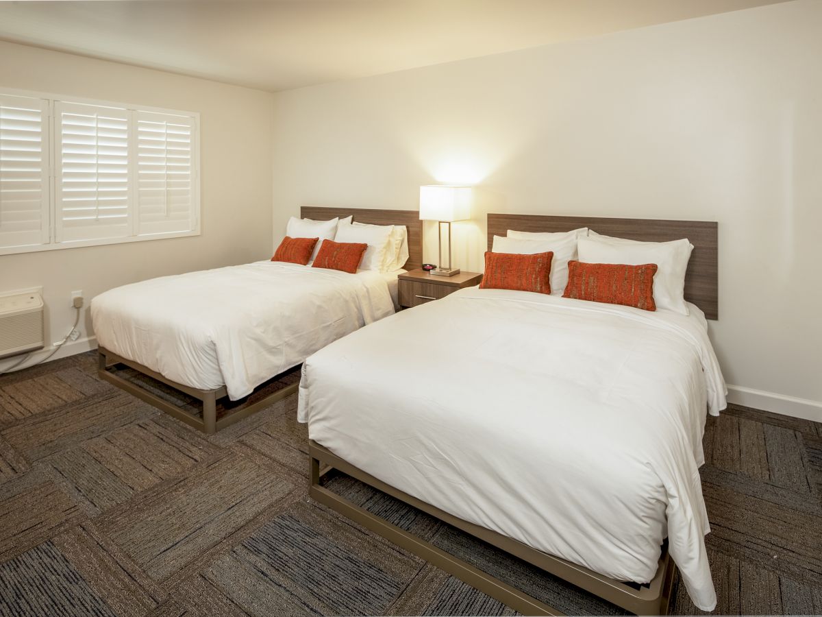 A minimalistic bedroom has two white beds with red pillows, a wooden nightstand with a lamp, white walls, and a window with shutters.