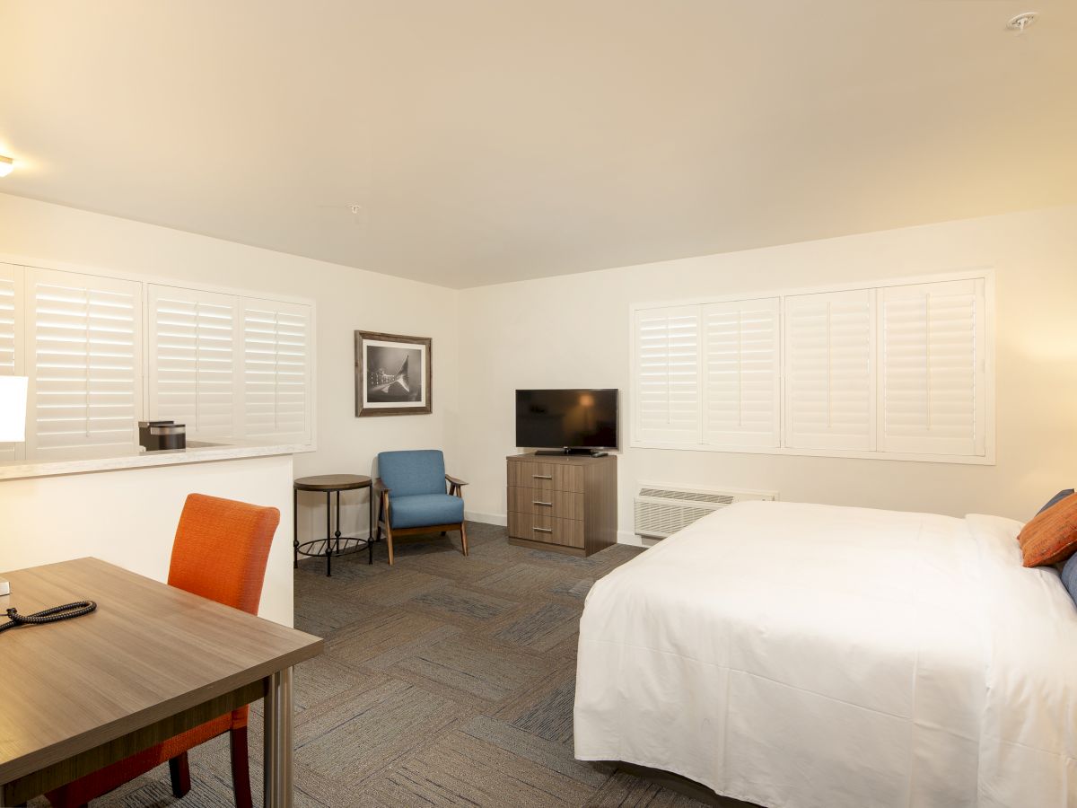 A spacious hotel room with a neatly made bed, desk, chairs, television, and artwork on the wall. The windows have white shutters, and there's soft lighting.