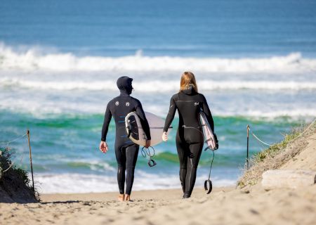 Two people in wetsuits carrying surfboards head towards a sandy beach with waves in the background.