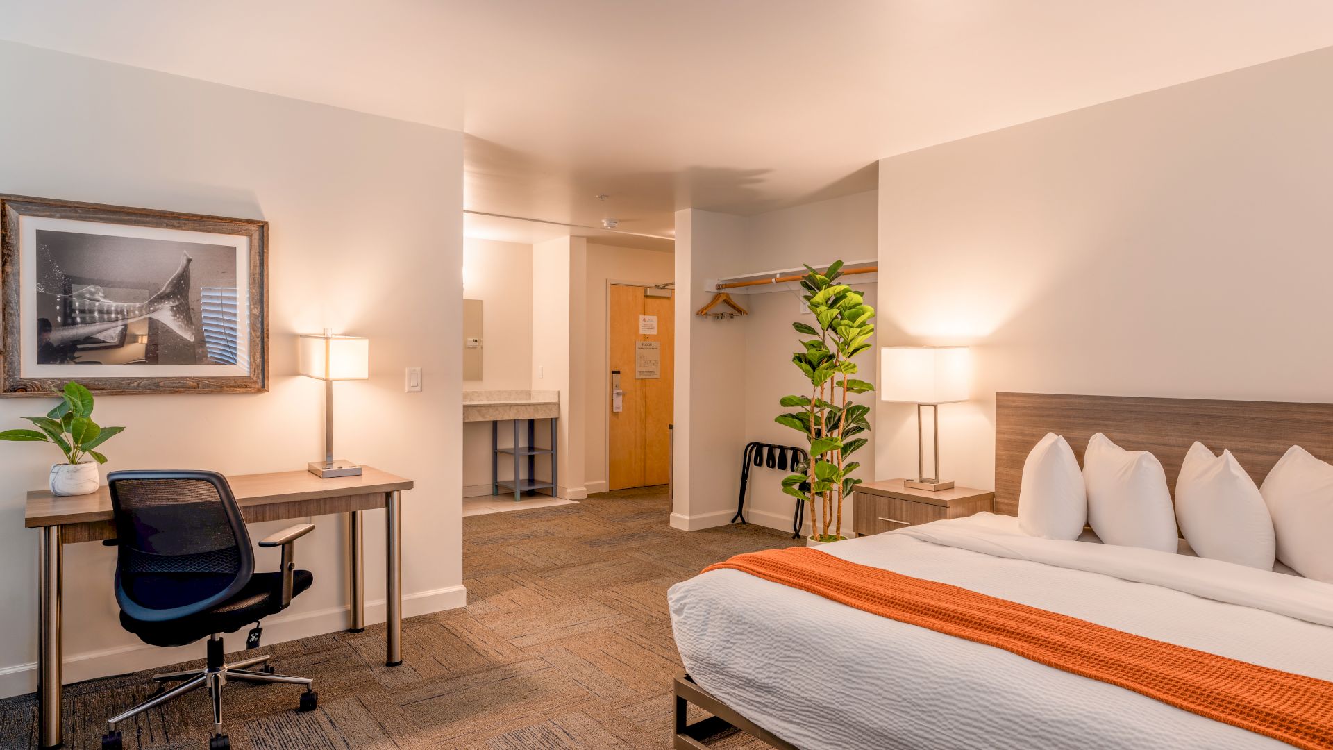 A hotel room with a bed, desk, chair, lamps, artwork, plant, open closet, and bathroom. The decor is modern and minimalist.