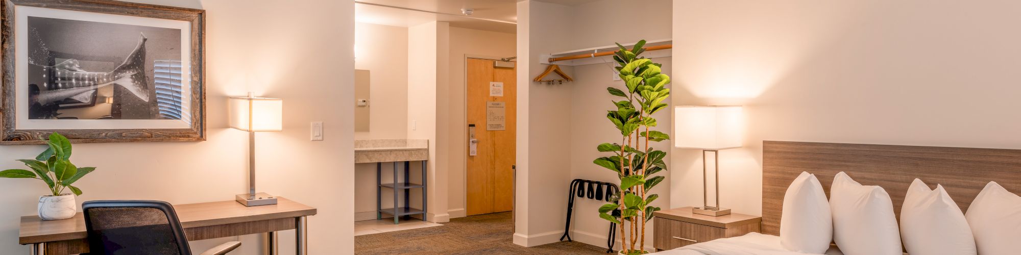 A hotel room with a bed, desk, chair, lamps, artwork, plant, open closet, and bathroom. The decor is modern and minimalist.