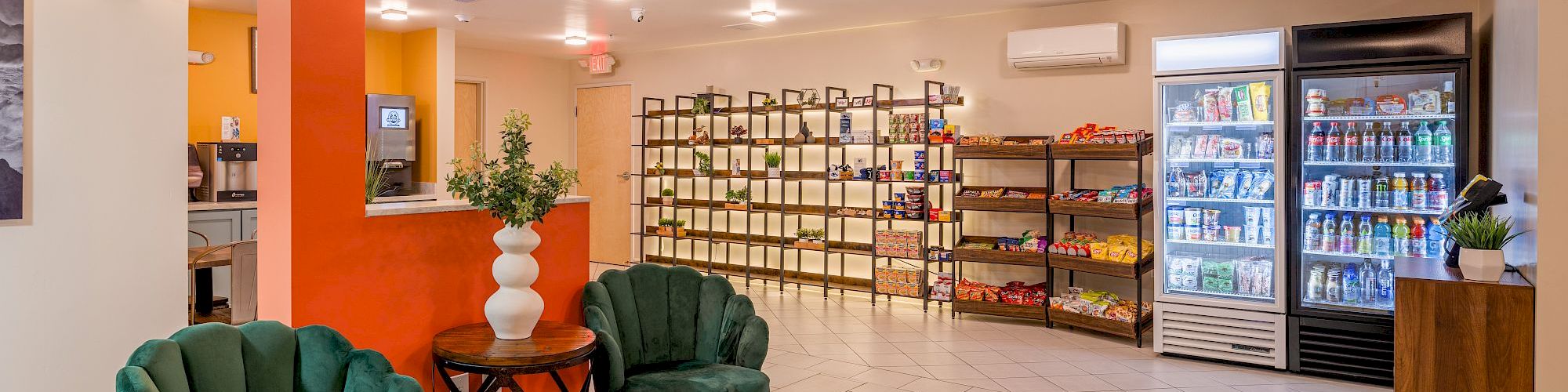 A well-lit room features green chairs, shelves with snacks, a fridge with drinks, and decorative plants on tables, creating a cozy atmosphere.