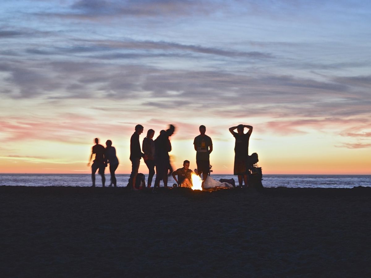 A group of people are gathered around a bonfire on a beach during sunset, silhouetted against the colorful sky.