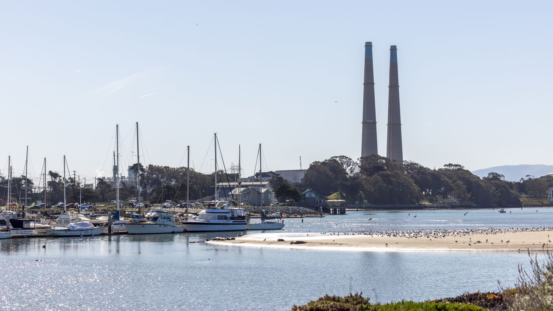The image shows boats docked in a marina with two tall smokestacks in the background, surrounded by calm water and clear skies.