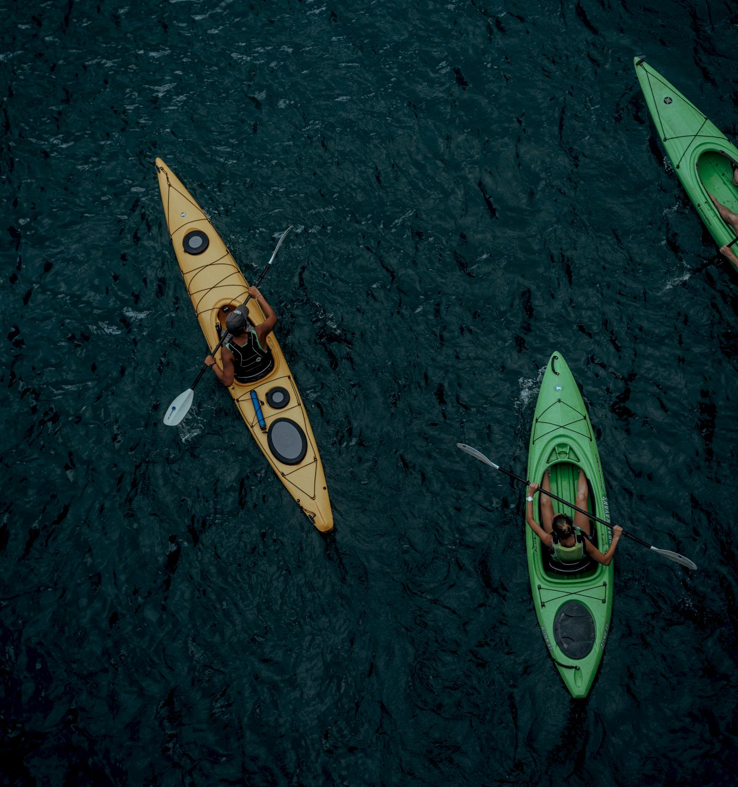 The image shows three kayakers paddling on dark, rippling water, using yellow and green kayaks viewed from an overhead angle.