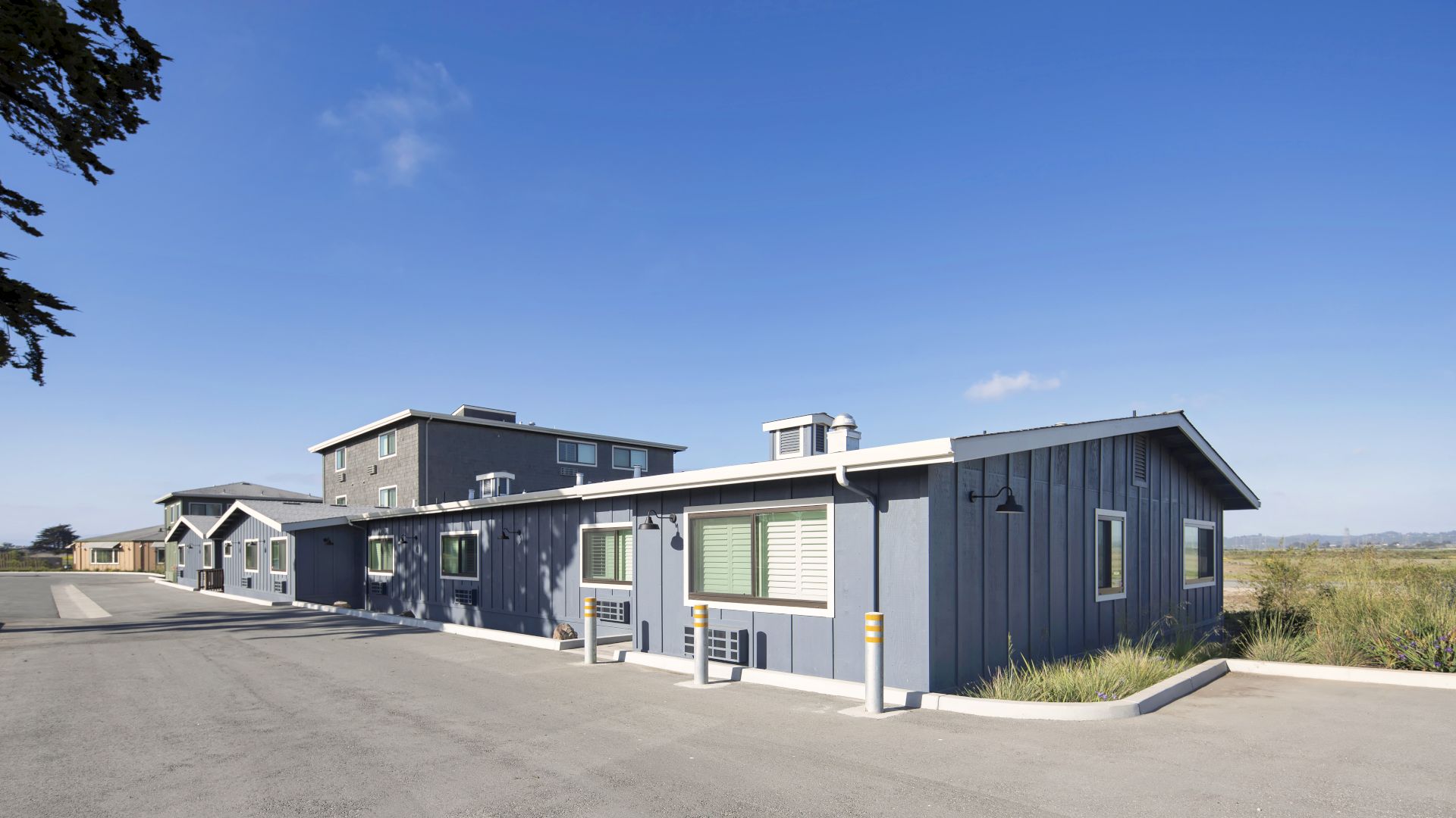 The image shows several modern, single-story buildings in a coastal setting with a clear blue sky in the background.