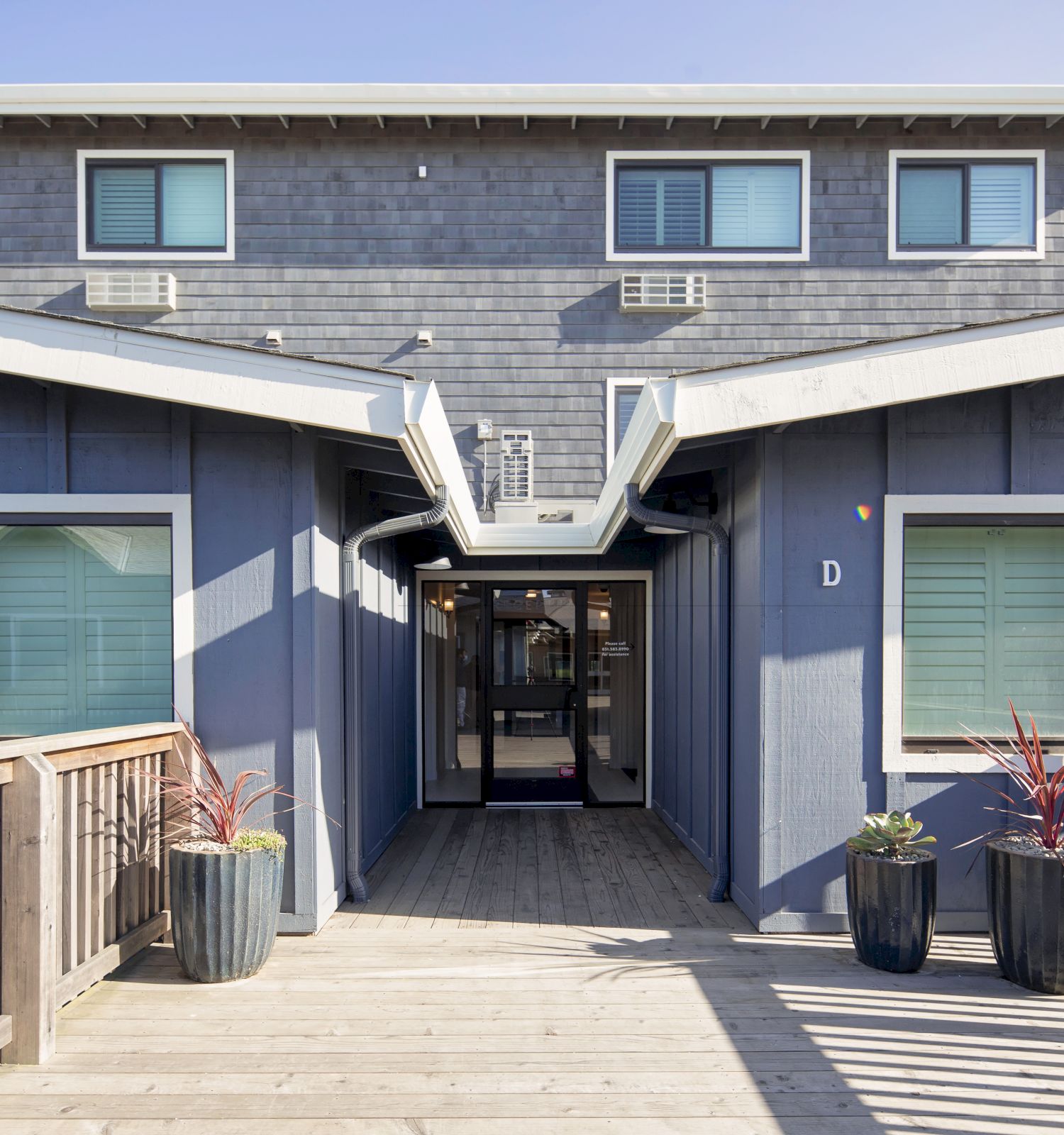 A modern building entrance with wooden ramps and railings, flanked by potted plants on either side, leading to a central doorway.