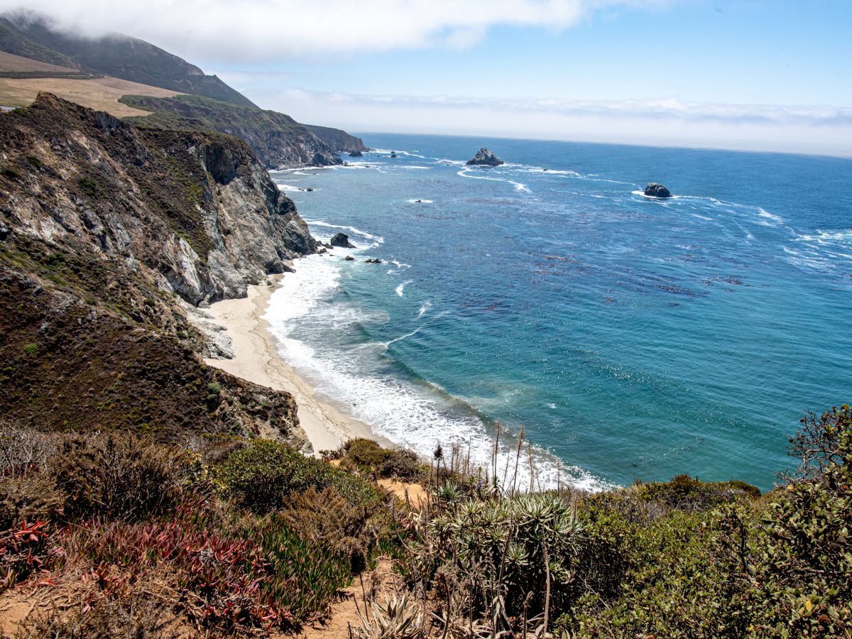 A scenic coastal view with rocky cliffs, a sandy beach, and the ocean with clear blue water and waves gently hitting the shore under a partly cloudy sky.