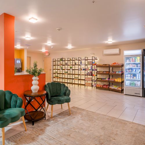 The image shows a bright room with green chairs, a table with a vase, snack shelves, and a fridge with drinks.