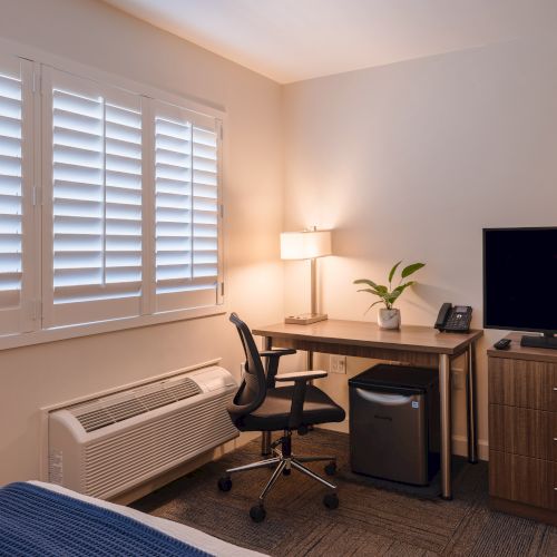 A modern room with a desk, chair, lamp, small fridge, TV stand, TV, and plant; shutters filter sunlight from the window.