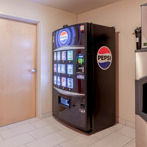 A room with a Pepsi vending machine, a light wood door, and an ice machine next to it.