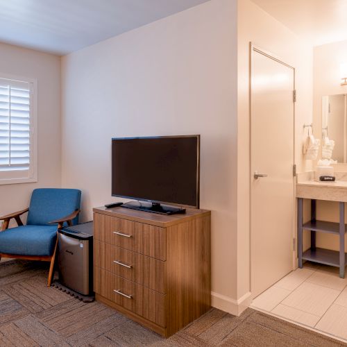 A hotel room features a TV on a wooden dresser, a blue chair, a small fridge, and a vanity sink area with a mirror and lighting.