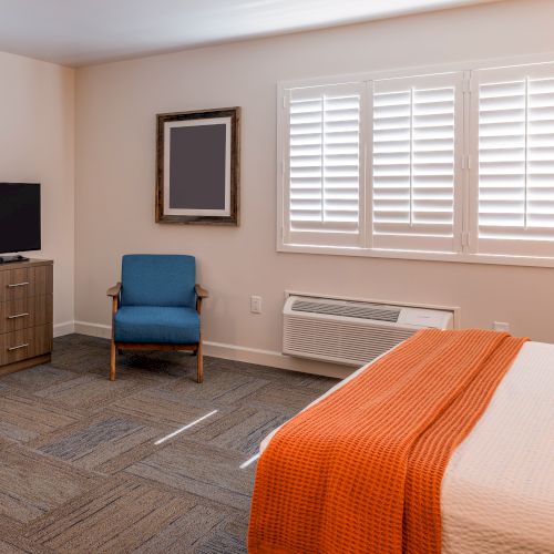 A hotel room with a bed, TV on a stand, blue chair, window with shutters, framed picture, and air conditioning unit on a carpeted floor.