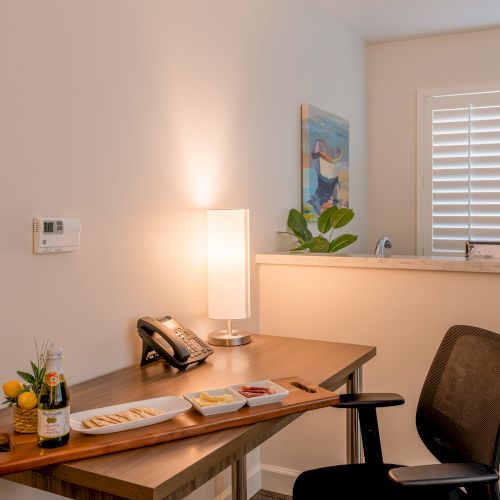 A cozy office desk with a phone, a lamp, snacks, and a small bottle, with a chair and window shutters visible.