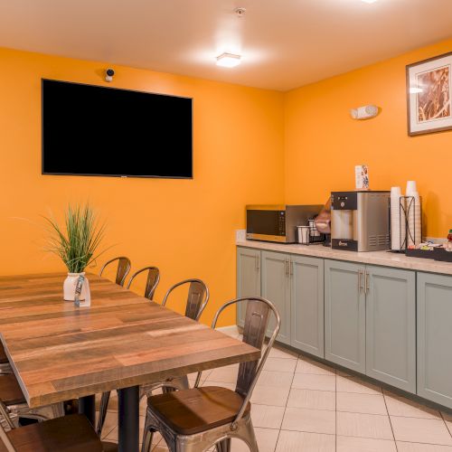 A brightly colored room with a wooden table, metal chairs, a TV, a plant, and a beverage machine on a counter.