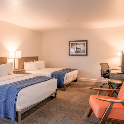 The image shows a hotel room with two beds, blue throws, a desk, chair, lamp, and a wall picture. There's also an orange armchair.