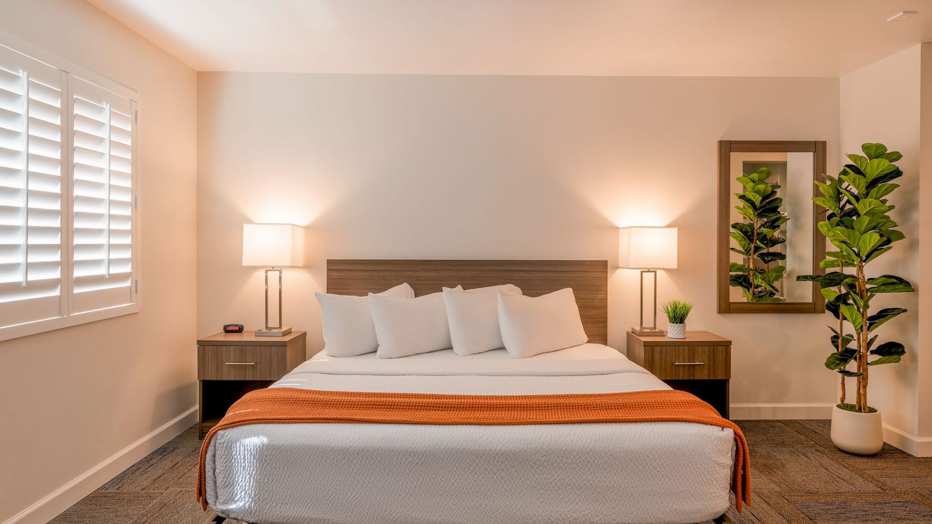 A neatly arranged bedroom featuring a large bed with white and orange bedding, two bedside tables with lamps, a mirror, and plants in the corner.