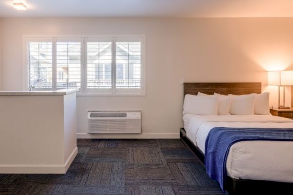 The image shows a hotel room with a bed, bedside lamp, window with shutters, air conditioning unit, and partial wall.