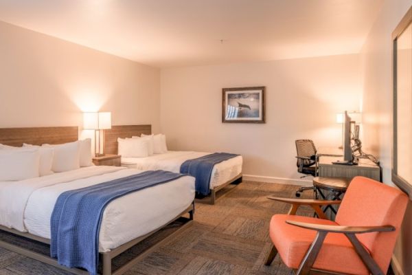 A hotel room with two double beds, a desk with chair, orange armchair, lamps, and artwork on the wall, looking clean and cozy.