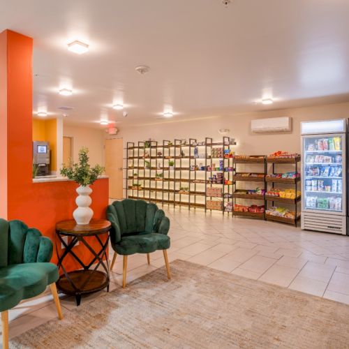 The image shows a lobby area with two green chairs, a small table, shelves with products, and a fridge with drinks.