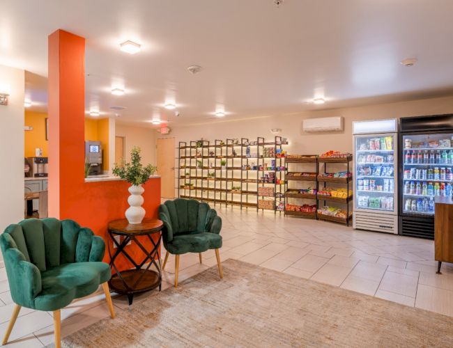 The image shows a lobby area with two green chairs, a small table, shelves with products, and a fridge with drinks.