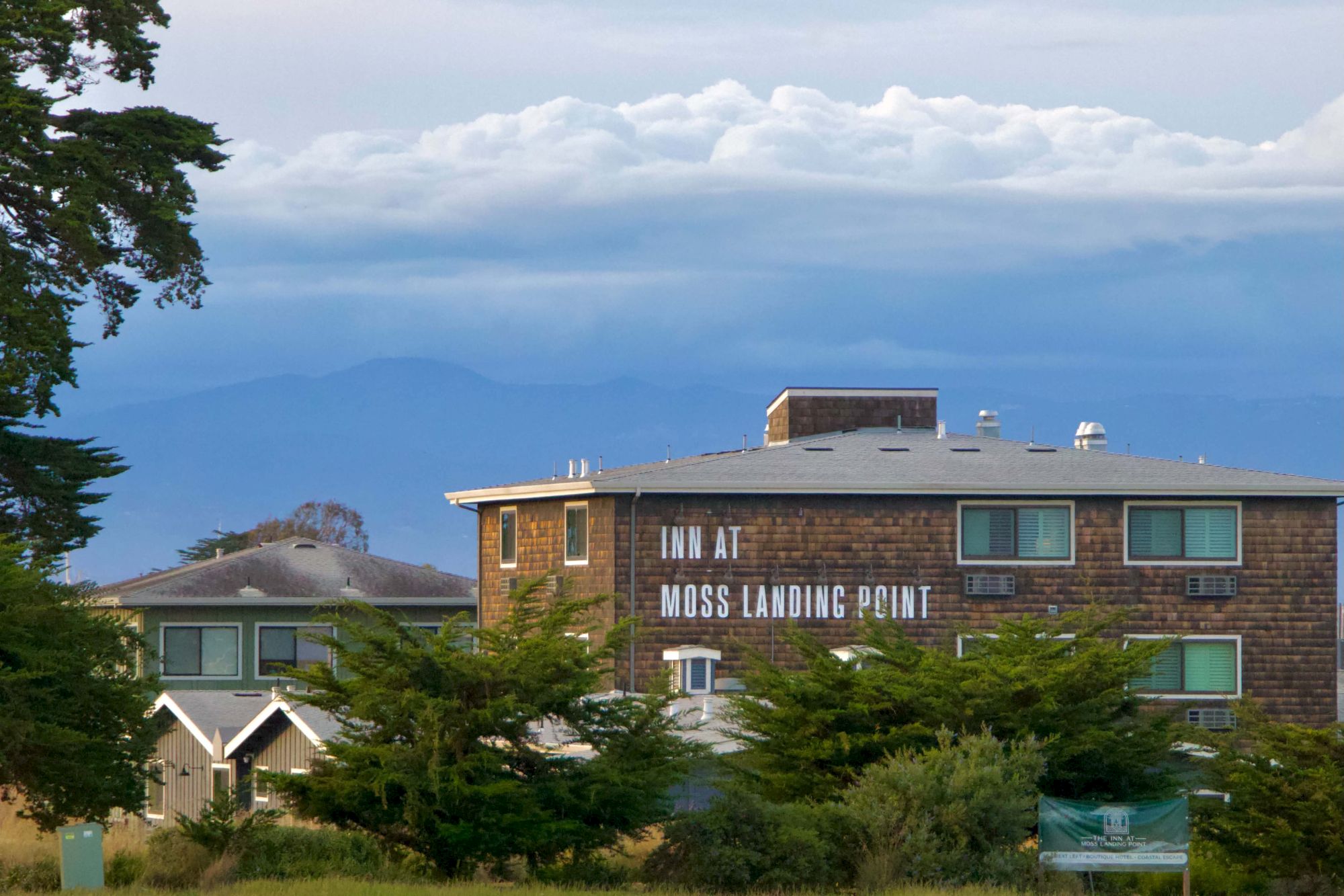 A brick building labeled &ldquo;Moss Landing Point&rdquo; sits among trees with hills and a cloudy sky in the background, a peaceful coastal town scene.