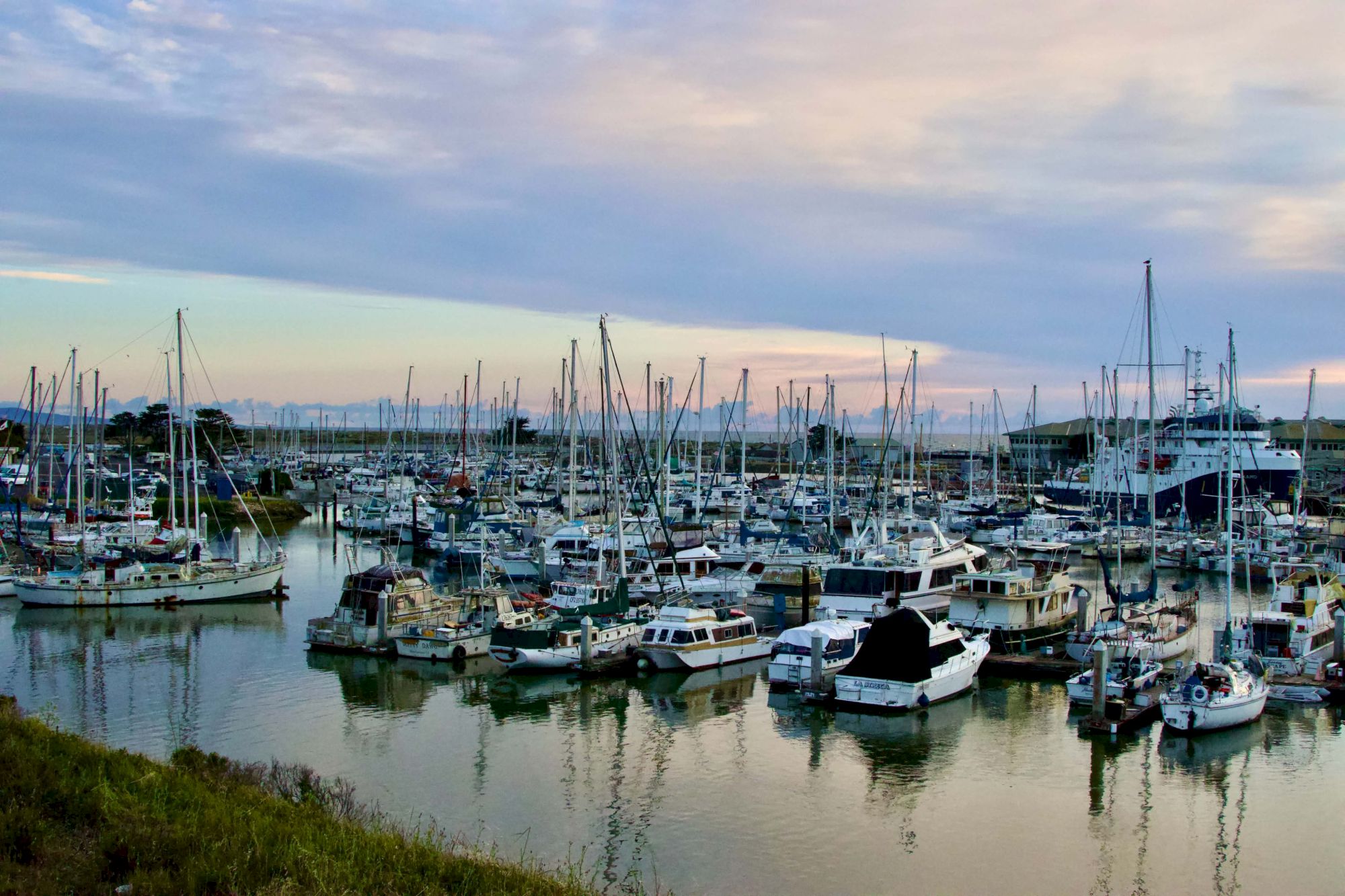 A harbor full of moored boats and yachts at sunset, calm water reflecting the masts, with a cloudy sky above.